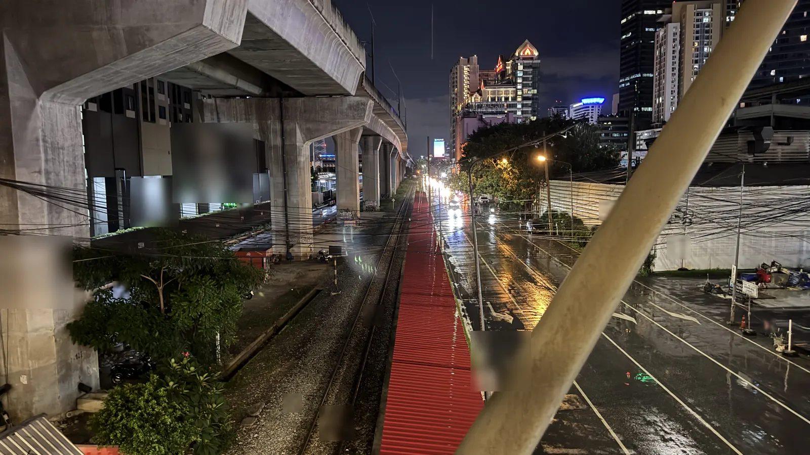 雨上がりの夜のバンコク。高架下を走る線路と、濡れた路面